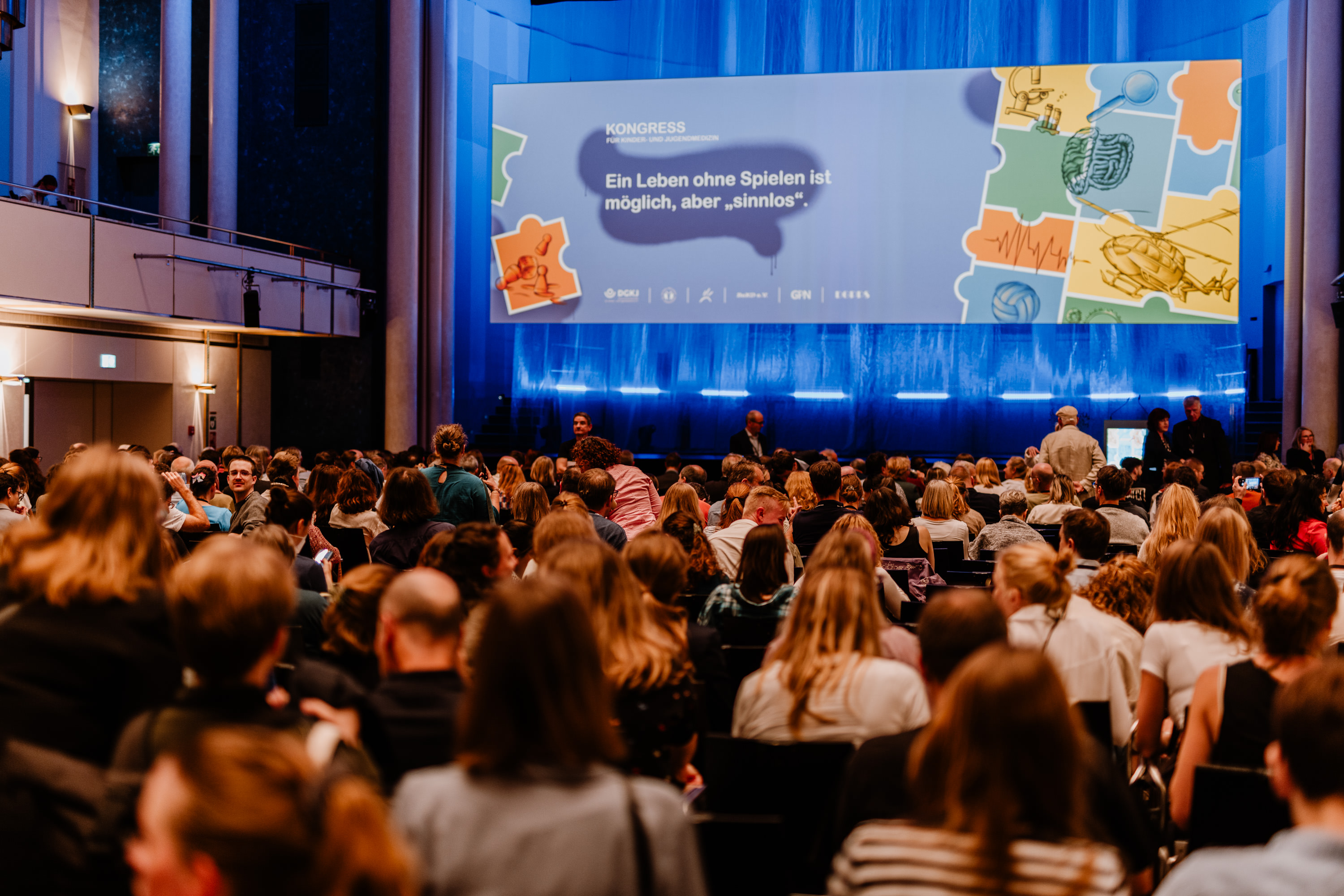 Großer Konferenzraum mit zahlreichen Teilnehmenden, die auf eine Bühne blicken. Auf der Bühne befindet sich eine große Leinwand mit der Aufschrift ‚Ein Leben ohne Spielen ist möglich, aber sinnlos‘, begleitet von bunten Illustrationen. Die Szene zeigt eine organisierte Veranstaltung oder Präsentation