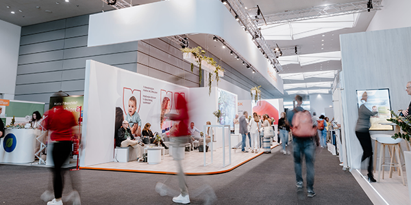Foto einer modernen Messehalle mit mehreren Ausstellungsständen und Besuchern. Menschen gehen umher oder sitzen in Gesprächsbereichen. Die Halle ist hell beleuchtet, mit weißen Wänden, Postern und Displays.