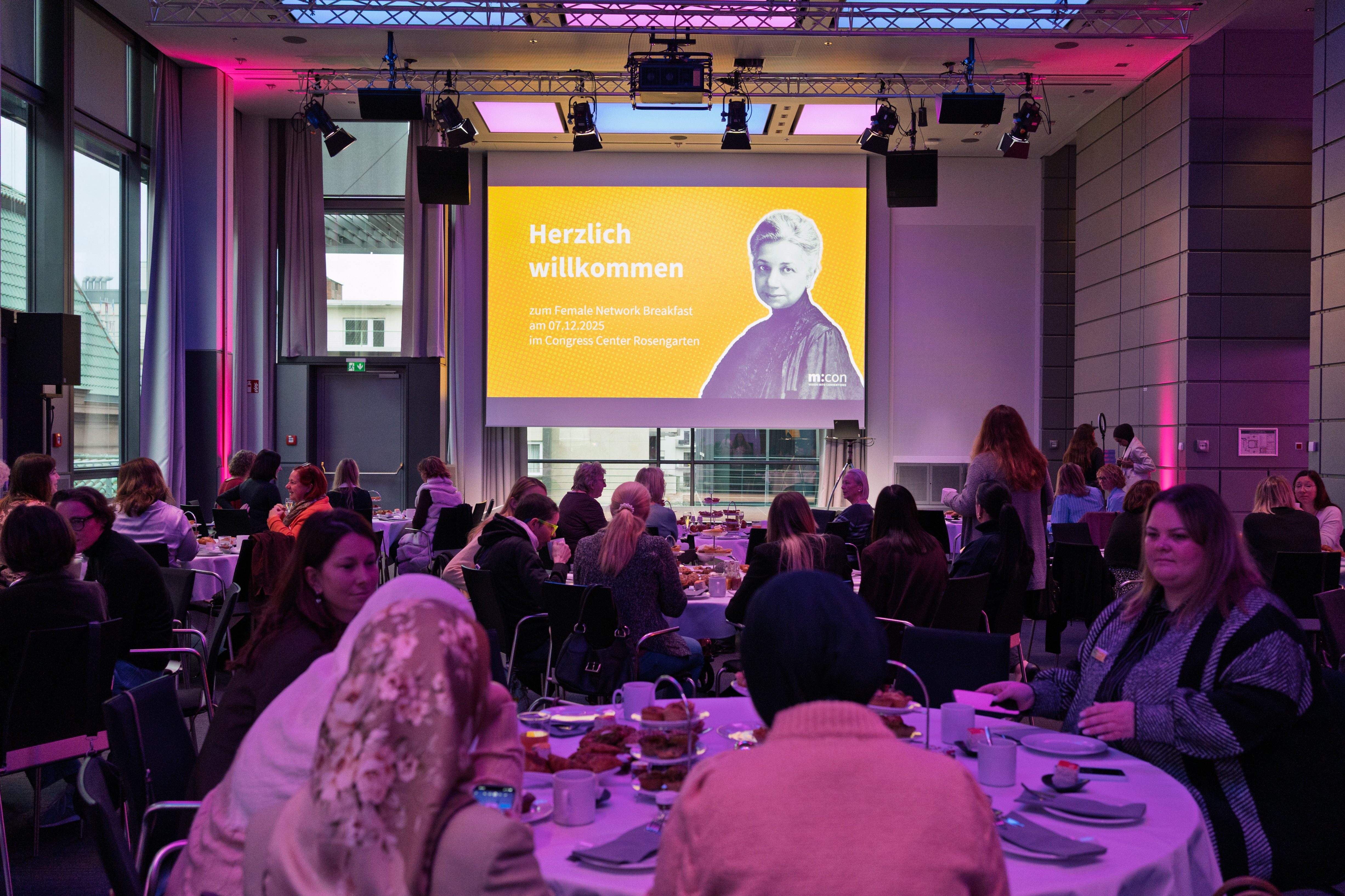 Ein Saal mit festlich gedeckten Tischen und Gästen, Leinwand auf der Bühne