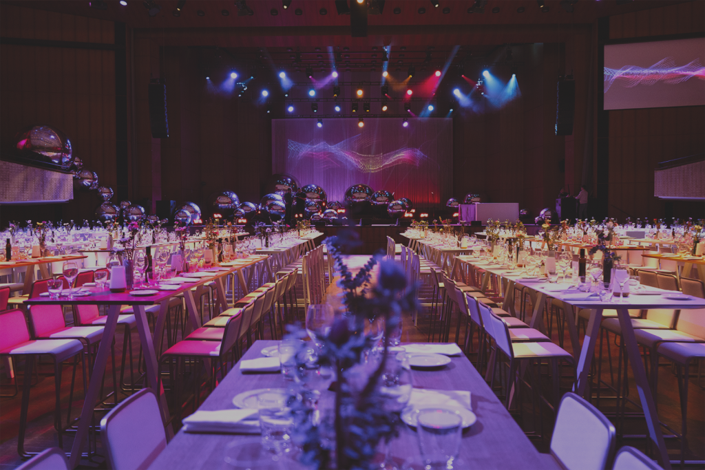 Großer Veranstaltungssaal mit mehreren langen, gedeckten Tischen und Stuhlreihen, dekoriert für ein Dinner‑Event; im Hintergrund eine Bühne mit Lichttechnik und großflächiger Projektion.