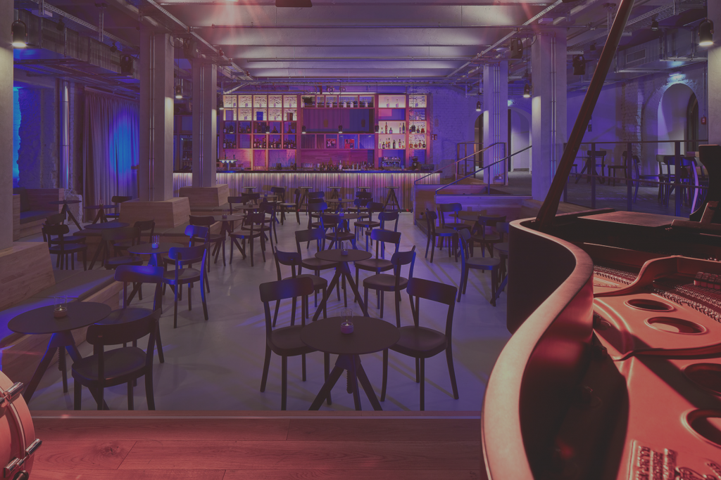 Innenraum einer modernen Jazzbar mit leerem Sitzbereich aus runden Tischen und Stühlen, einer erhöhten Bühne und einer beleuchteten Bar im Hintergrund; im Vordergrund ist ein Flügel zu sehen, das Licht ist in Blau‑ und Violetttönen gehalten.