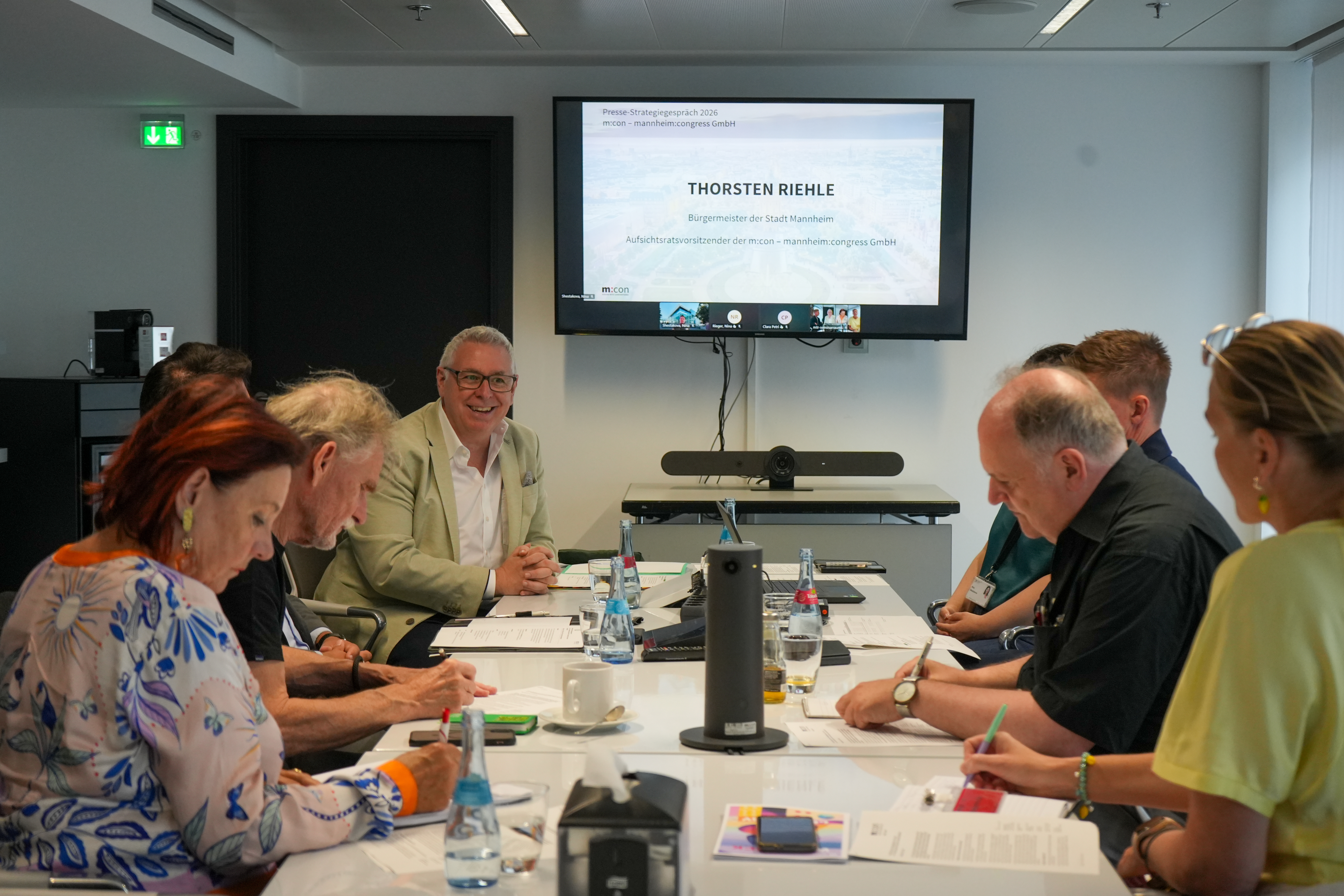 Männer und Frauen sitzen an einem Tisch bei einer Präsentation mit einem Bildschirm im Hintergrund